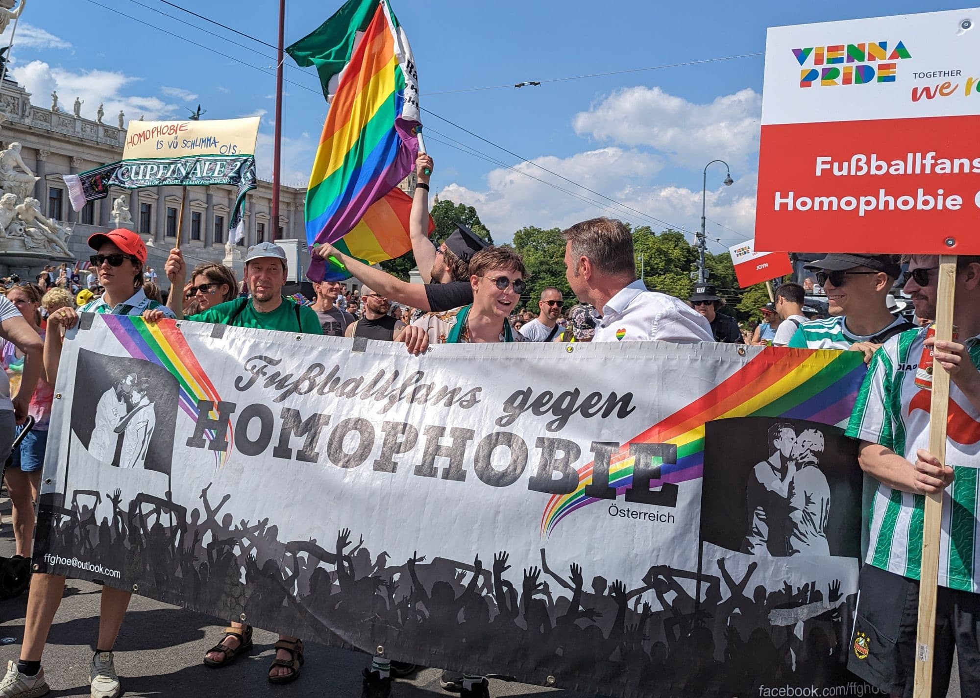 Queere Fußballfans auf der Wiener Regenbogenparade lachen hinter einem Banner gegen Homophobie im Fußball. Im Hintergrund, das österreichische Parlament.